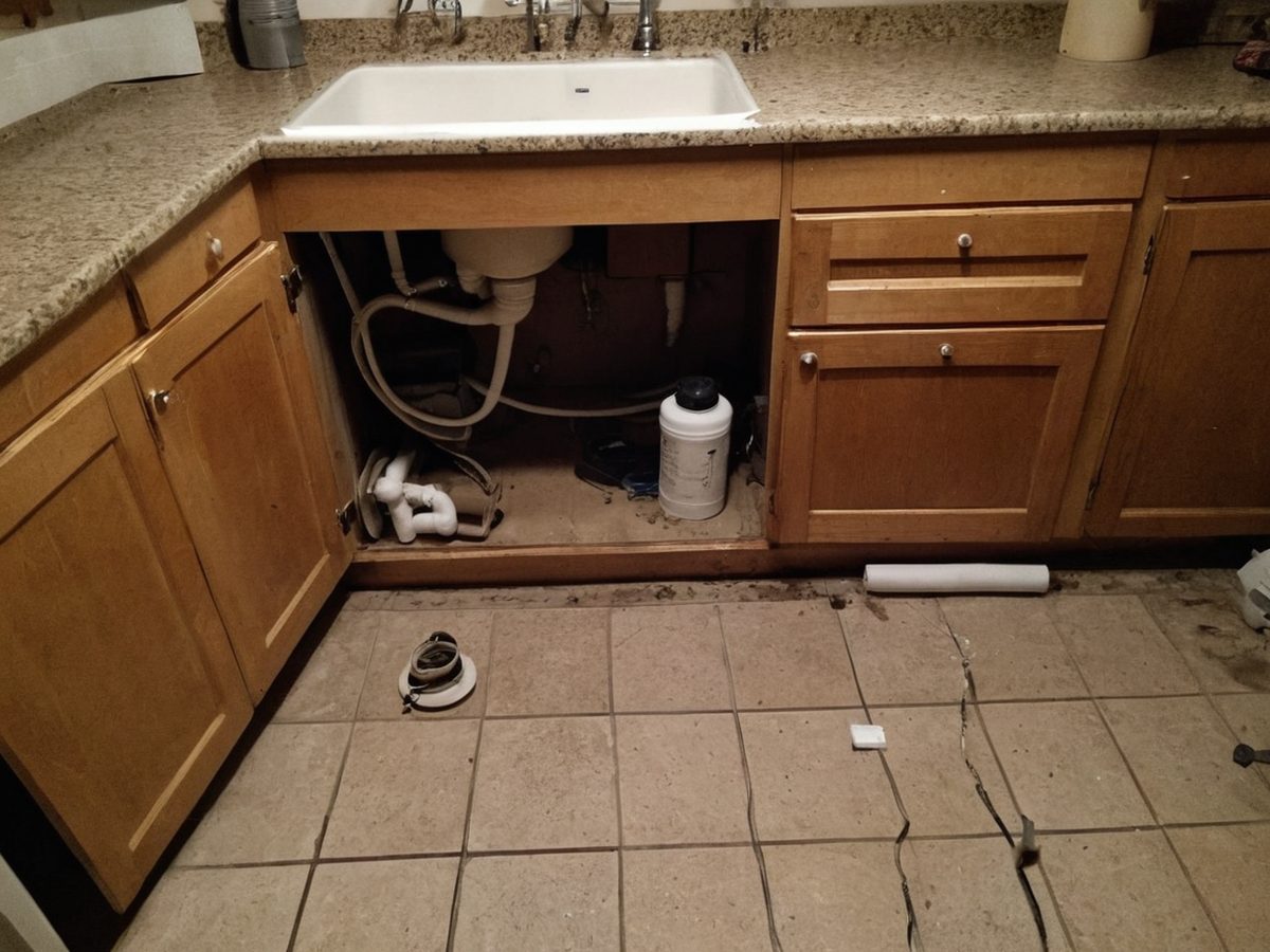 Open kitchen sink cabinet with PVC drain piping and a drain auger cable on the floor